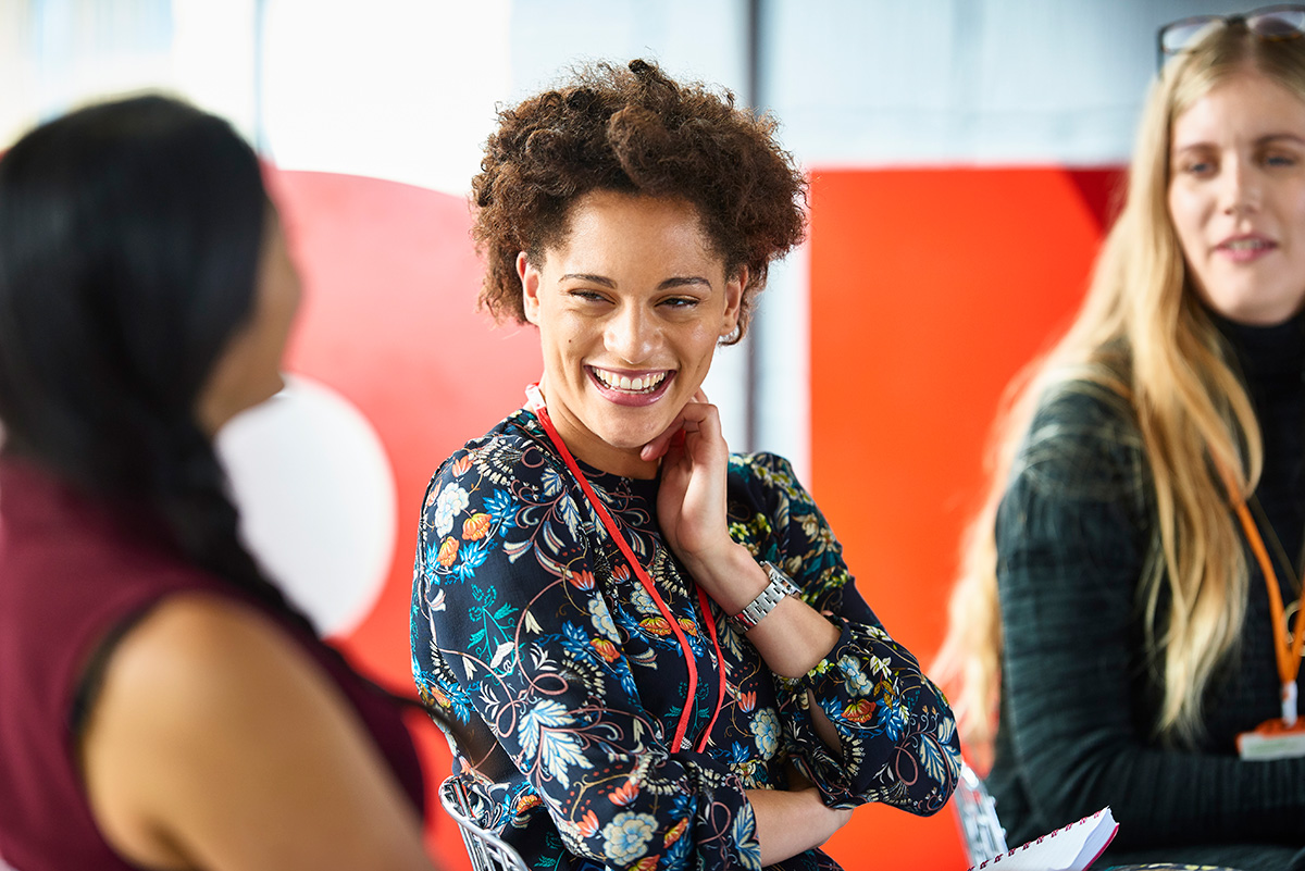 Stock photo of a smiling woman of color, two other woman are in frame but out of focus.  They look like they are at a conference.